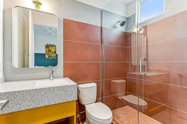 Contemporary bathroom interior with glass-enclosed shower and rainfall showerhead, large terracotta wall tiles and mosaic floor, white toilet, speckled countertop sink on a yellow vanity and wall mirror.