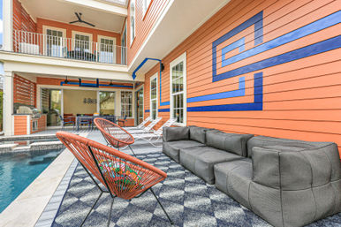 Coral-colored poolside patio with bold blue geometric mural, gray modular sofa, orange woven lounge chairs, white chaise lounges, outdoor dining area and second-floor balcony.
