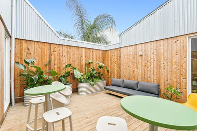 Bright urban outdoor patio with wooden decking and tall timber fence, green round tables with white stools, a gray minimalist sofa, corrugated metal planter boxes filled with tropical plants and palm fronds under a clear blue sky.