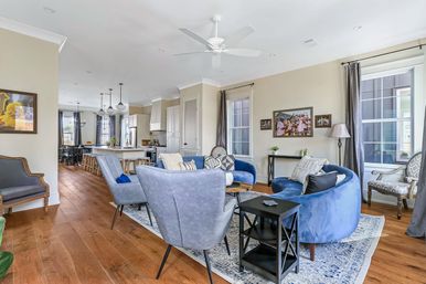 Bright open-concept living room and kitchen — blue velvet curved sofa, gray accent chairs, hardwood floors, kitchen island with stools, large windows and ceiling fan