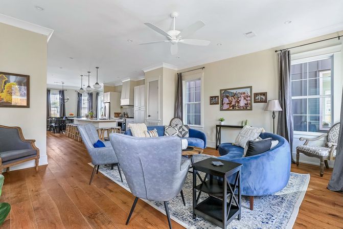 Bright open-concept living room and kitchen — blue velvet curved sofa, gray accent chairs, hardwood floors, kitchen island with stools, large windows and ceiling fan