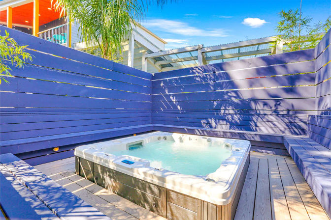 Inviting outdoor hot tub bubbling on a sunlit wooden deck, enclosed by a navy-blue slatted privacy fence with built-in bench seating, palm shadows and bright blue sky overhead.