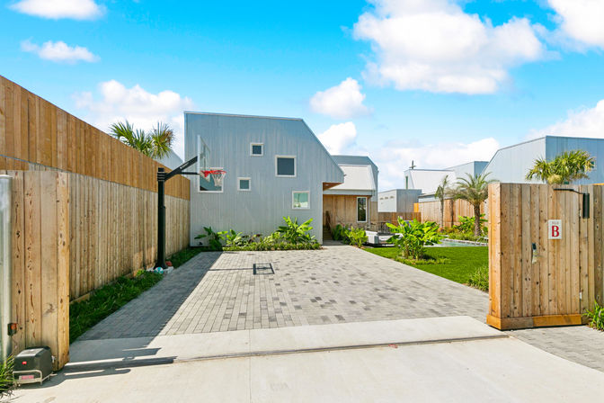 Gated modern villa courtyard with corrugated metal façade, paved driveway and basketball hoop, tropical plants and palm trees under a bright blue sky.