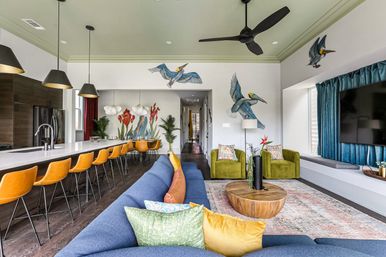 Vibrant open-plan living room and kitchen with a long white island lined by orange bar stools, a blue sectional with colorful pillows, green armchairs, round wooden coffee table, ceiling fan, large TV, and tropical bird and plant wall murals.