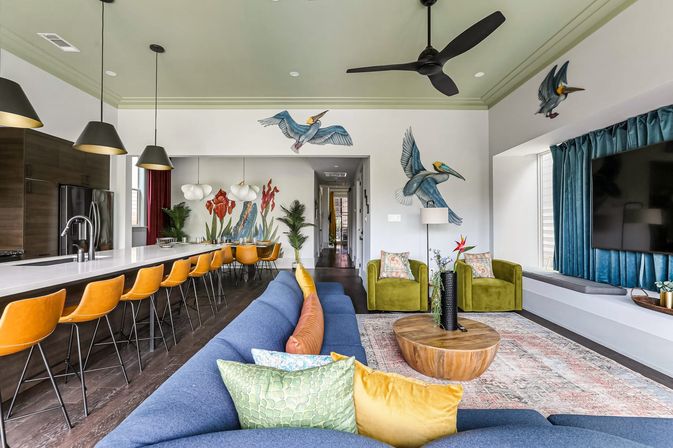 Vibrant open-plan living room and kitchen with a long white island lined by orange bar stools, a blue sectional with colorful pillows, green armchairs, round wooden coffee table, ceiling fan, large TV, and tropical bird and plant wall murals.