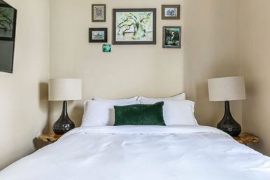 Cozy bedroom with white bedding and duvet, emerald green accent pillow, dark ceramic bedside lamps on live-edge wooden nightstands, and a small gallery wall of nature artwork on neutral walls.