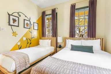 Bright modern guest room with a queen and twin bed, wooden slat headboards, white linens and patterned throws, yellow floral wall mural with framed art, tall windows with purple curtains and a tree-lined view.