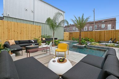 Sunny urban backyard patio with rectangular plunge pool, black modular sofas, yellow accent chair, palm trees, wood privacy fence and tiled deck — modern outdoor lounge