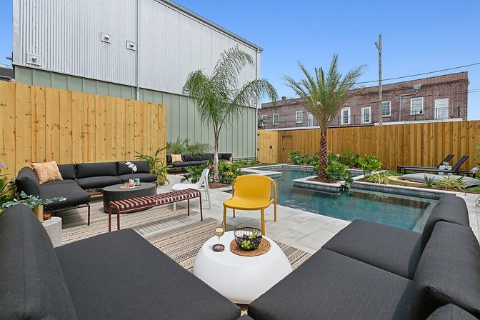 Sunny urban backyard patio with rectangular plunge pool, black modular sofas, yellow accent chair, palm trees, wood privacy fence and tiled deck — modern outdoor lounge