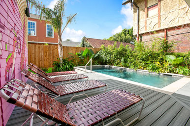 Sunny urban backyard with a rectangular swimming pool, gray wood deck, row of woven maroon lounge chairs, pink mural on the siding, palm tree and tropical plants along a wooden fence