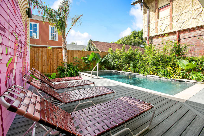 Sunny urban backyard with a rectangular swimming pool, gray wood deck, row of woven maroon lounge chairs, pink mural on the siding, palm tree and tropical plants along a wooden fence