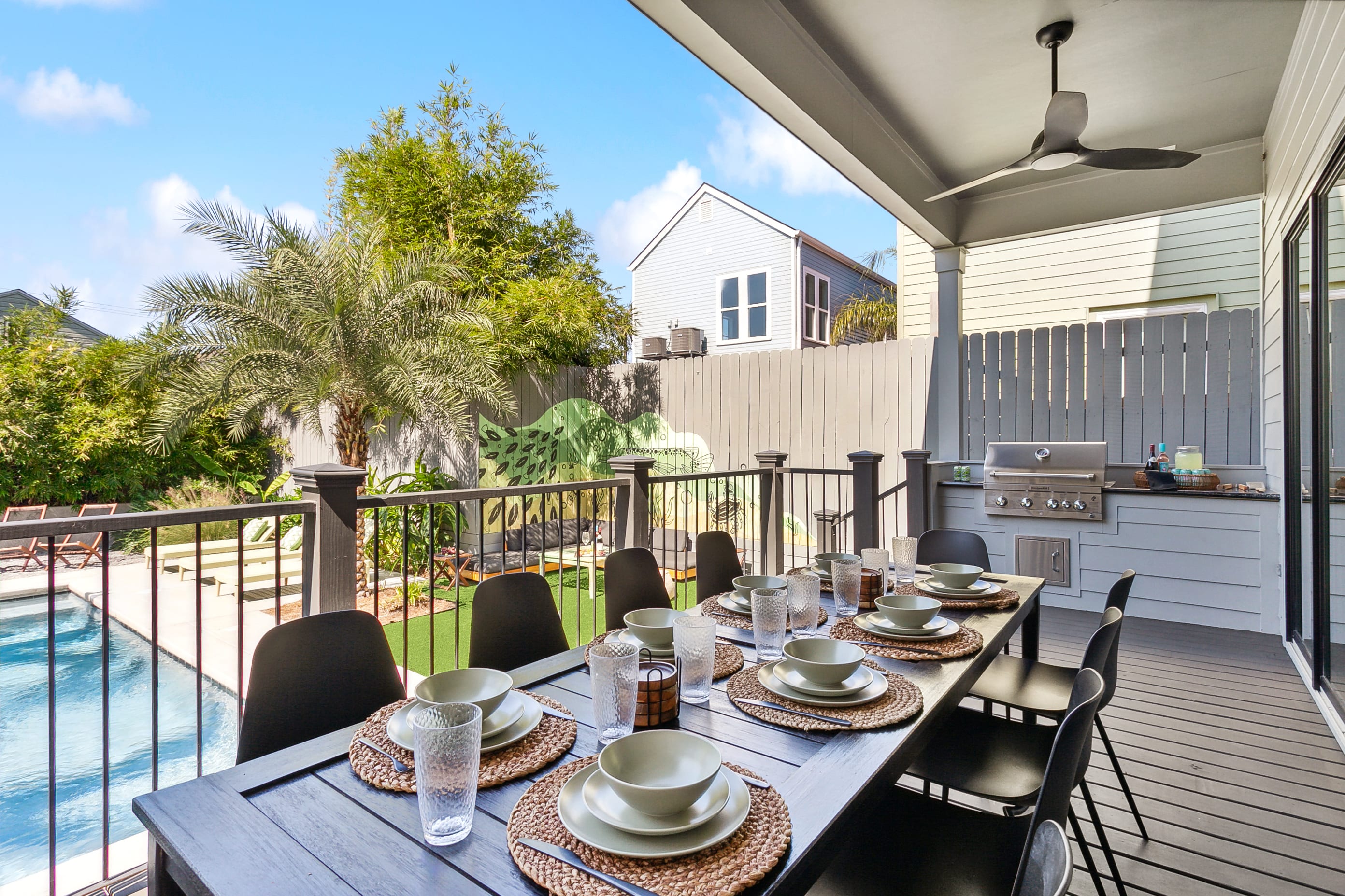 Covered outdoor dining patio set for eight overlooking a pool and palm-tree backyard with built-in stainless grill and ceiling fan