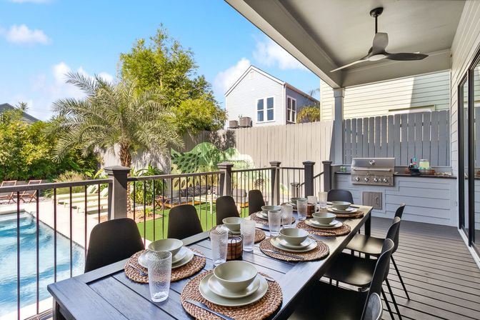 Covered outdoor dining patio set for eight overlooking a pool and palm-tree backyard with built-in stainless grill and ceiling fan