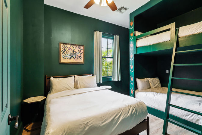 Cozy bedroom with deep green walls, queen bed dressed in white linens, stacked wood bunk beds with hand-painted floral post and ladder, window with light curtains and ceiling fan