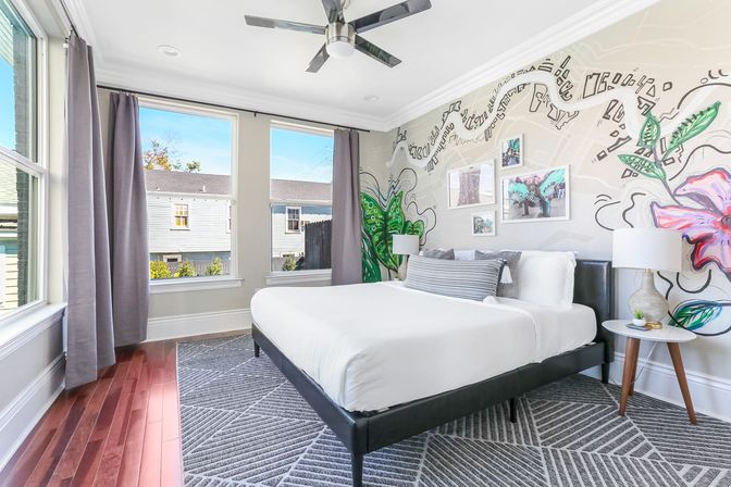 Sunlit modern bedroom in an urban home with a white-dressed bed and gray accent pillow, geometric area rug over hardwood floors, large windows with gray curtains, ceiling fan, bedside tables with lamps, and a colorful floral and abstract wall mural.