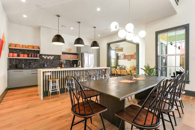 Bright, light-filled open-concept kitchen and dining area with a long dark wood table and black spindle chairs, adjacent island with bar stools and three black pendant lights, stainless fridge, open shelves with orange dishes, and wide-plank hardwood floors.