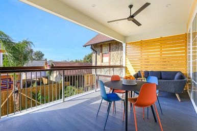 Sunny covered balcony patio with modern round dining table and four colorful chairs, wicker lounge sofa, ceiling fan, wooden privacy screen and neighborhood rooftop views