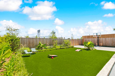 Sunny residential backyard with bright artificial turf, two lounge chairs, cornhole boards, landscaped planting beds, paved patio and a basketball hoop against a wooden privacy fence under a blue sky