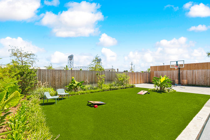 Sunny residential backyard with bright artificial turf, two lounge chairs, cornhole boards, landscaped planting beds, paved patio and a basketball hoop against a wooden privacy fence under a blue sky