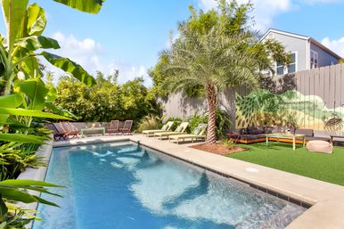 Sunny tropical backyard pool with palm tree and banana plants, green chaise lounges, mural-painted fence and relaxed outdoor seating area