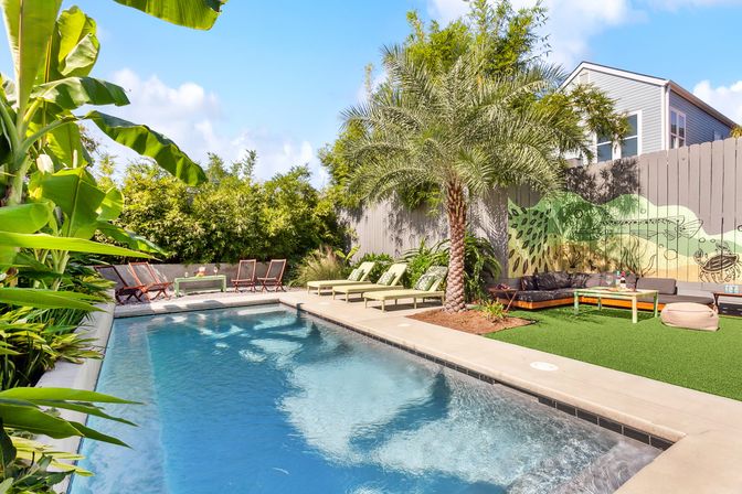 Sunny tropical backyard pool with palm tree and banana plants, green chaise lounges, mural-painted fence and relaxed outdoor seating area