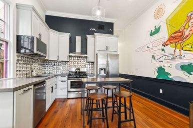 Contemporary kitchen with white cabinets, stainless steel appliances, geometric tile backsplash, wooden island with four stools, warm hardwood floors and a colorful bird mural