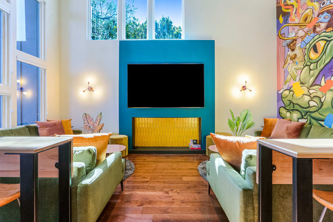 Vibrant modern living room with green velvet sofas, teal TV wall and golden panel, colorful mural, large windows and hardwood floors.