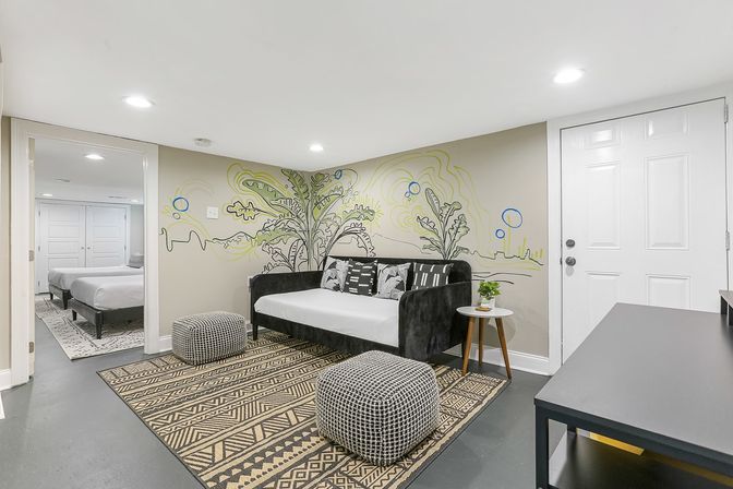 Cozy lower-level lounge with black daybed, botanical wall mural, geometric rug and two patterned poufs; round side table with plant, recessed lighting and open doorway into a bedroom with two beds.