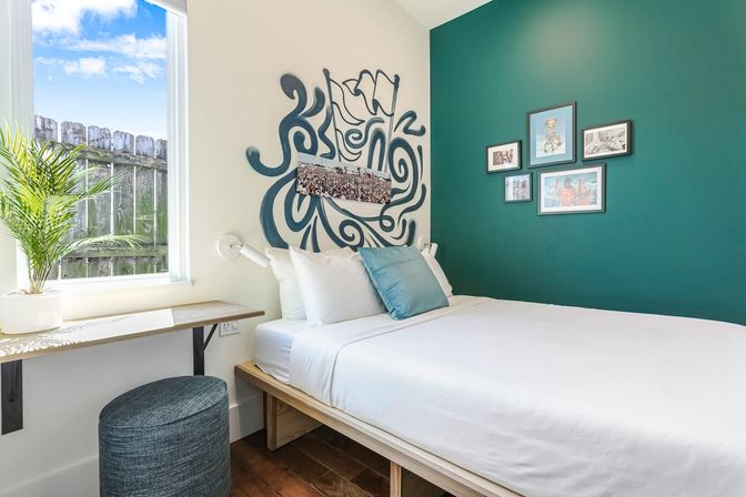 Bright, cozy modern bedroom with natural light, white linens on a low wooden platform bed, teal accent wall with framed artwork and abstract mural, window desk with potted plant and a textured ottoman.