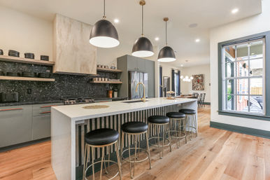 Bright modern open‑concept kitchen with a long white marble island, five black leather bar stools, matte black pendant lights, stainless refrigerator, dark speckled backsplash and light hardwood floors — stylish entertaining space with adjacent dining area and large window.