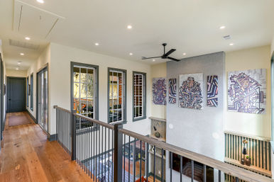 Bright upstairs hallway in a modern loft-style home with warm hardwood floors, black metal railing overlooking a double-height living area, geometric canvas art, gray-trimmed windows, recessed lights and a black ceiling fan.