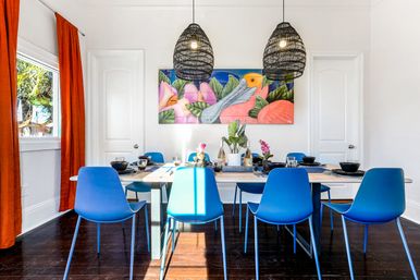 Bright coastal dining room with a wooden table set for six and vibrant blue chairs, woven black pendant lights, colorful tropical bird wall art, orange curtains, white walls and dark hardwood floors.