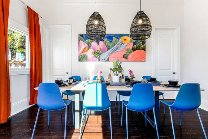 Bright coastal dining room with a wooden table set for six and vibrant blue chairs, woven black pendant lights, colorful tropical bird wall art, orange curtains, white walls and dark hardwood floors.