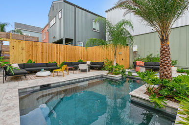 Urban backyard pool oasis with modern gray townhouses, wooden fence, palm trees, black sectional seating and colorful chairs on a tiled patio.
