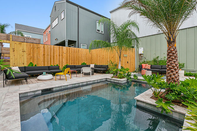 Urban backyard pool oasis with modern gray townhouses, wooden fence, palm trees, black sectional seating and colorful chairs on a tiled patio.