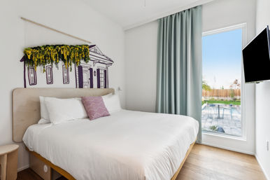 Bright modern bedroom with white linens and wooden headboard, purple accent pillow, wall art of a house facade topped with hanging greenery, pale green floor-to-ceiling curtain, large window with backyard patio and swimming pool view, and a wall-mounted TV.