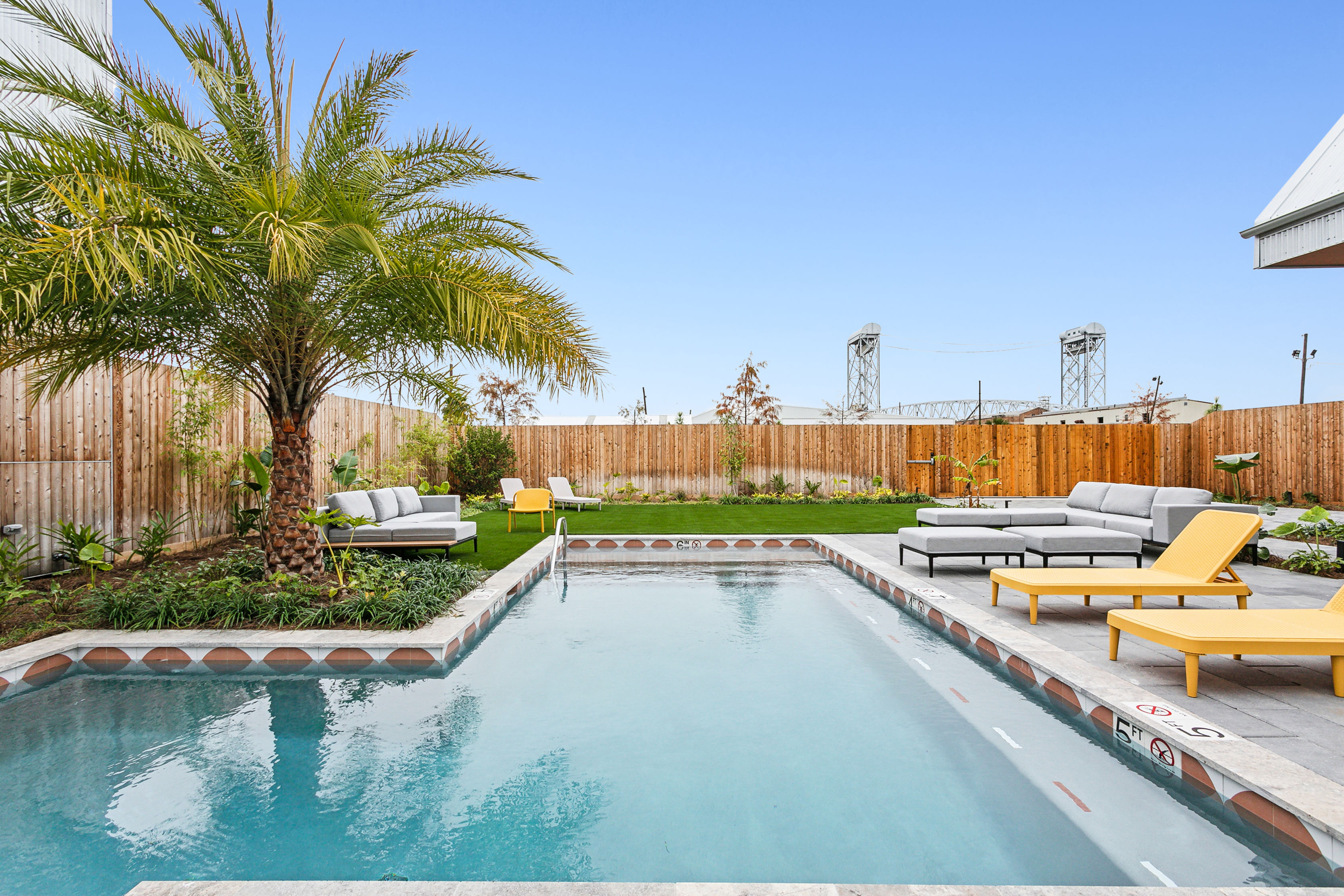 Sunlit residential backyard with a rectangular swimming pool, large palm tree, gray outdoor sectional and bright yellow lounge chairs on a modern patio, green lawn and wooden fence under a clear blue sky.
