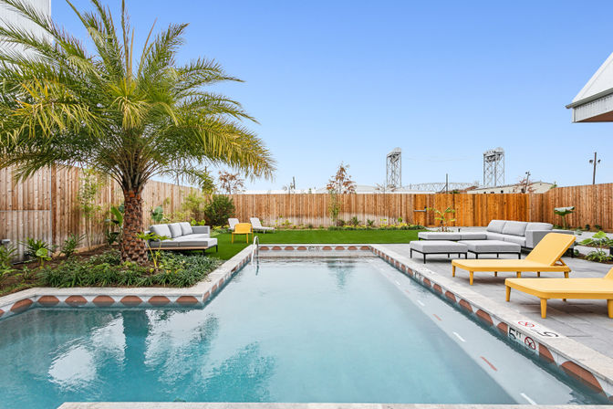 Sunlit residential backyard with a rectangular swimming pool, large palm tree, gray outdoor sectional and bright yellow lounge chairs on a modern patio, green lawn and wooden fence under a clear blue sky.