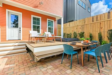 Bright backyard patio with coral-orange siding, raised wooden deck with white lounge chairs and a black hot tub, round wooden dining table set with teal chairs on a brick paver patio beside a tall wooden privacy fence.