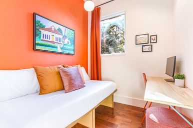 Cozy sunlit bedroom with orange accent wall, white daybed topped with rust and mauve pillows, wooden desk with monitor, orange curtains, framed art and hardwood floor.