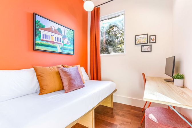 Cozy sunlit bedroom with orange accent wall, white daybed topped with rust and mauve pillows, wooden desk with monitor, orange curtains, framed art and hardwood floor.