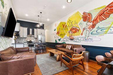 Open-plan loft-style living room and kitchen with a playful flamingo wall mural, hardwood floors, brown leather sofas, wooden coffee table, mid-century accent chairs, gray area rug, stainless steel appliances and a breakfast island.