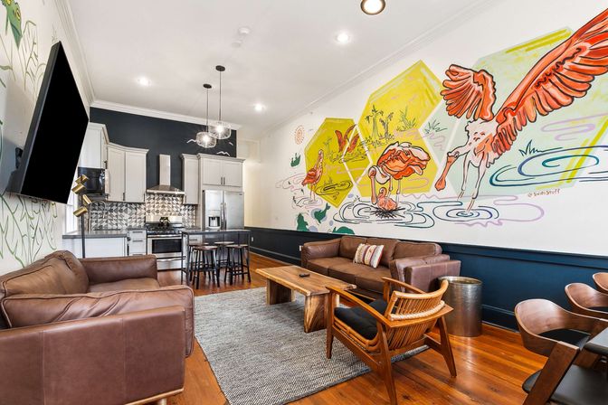 Open-plan loft-style living room and kitchen with a playful flamingo wall mural, hardwood floors, brown leather sofas, wooden coffee table, mid-century accent chairs, gray area rug, stainless steel appliances and a breakfast island.