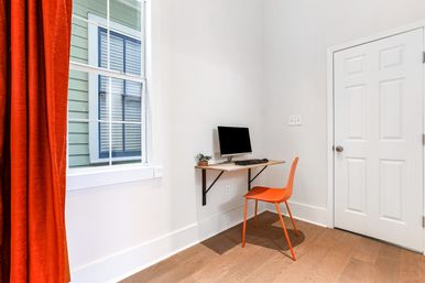 Minimalist home office nook with a floating wooden desk holding a monitor, keyboard and small plant, bright orange chair, red-orange curtain by a sunlit window and light hardwood floor.