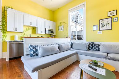 Sunny open-plan kitchen and living nook with bright yellow walls, white cabinets and stainless appliances, large window, built-in L-shaped gray banquette with geometric pillows, hardwood floors and small coffee tables