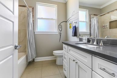 Bright modern home bathroom with white shaker vanity and dark quartz countertop, chrome faucets, toilet beneath frosted window, striped shower curtain over tub, neutral tile floor and playful line-art wall decal