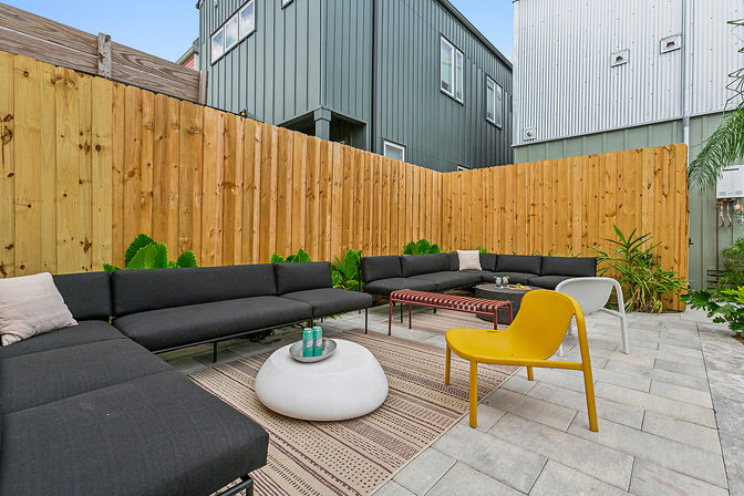 Modern urban backyard patio with L-shaped dark gray sectional sofa, yellow accent chair, white round stone coffee table, wooden privacy fence, paver flooring and green plants — stylish outdoor lounge