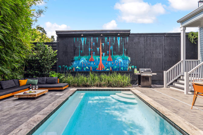 Residential backyard with a rectangular swimming pool and tiled steps, gray paver patio, L-shaped outdoor sofa with colorful cushions and a low wooden coffee table with wine glasses, stainless steel grill by house stairs, and a vibrant turquoise-orange nature mural on a black fence.