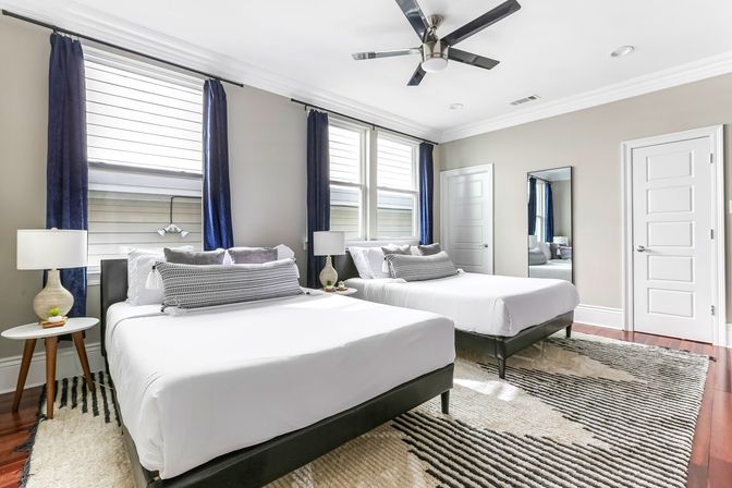 Bright modern guest bedroom with two neatly made beds, navy curtains framing double windows, mid-century nightstands with lamps, black ceiling fan, full-length mirror, patterned area rug, and hardwood floors
