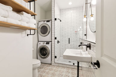 Bright modern bathroom with stacked washer and dryer beside a glass-enclosed subway-tile shower, wall-mounted sink, open wood shelving with folded white towels, patterned floor tile and matte-black fixtures.